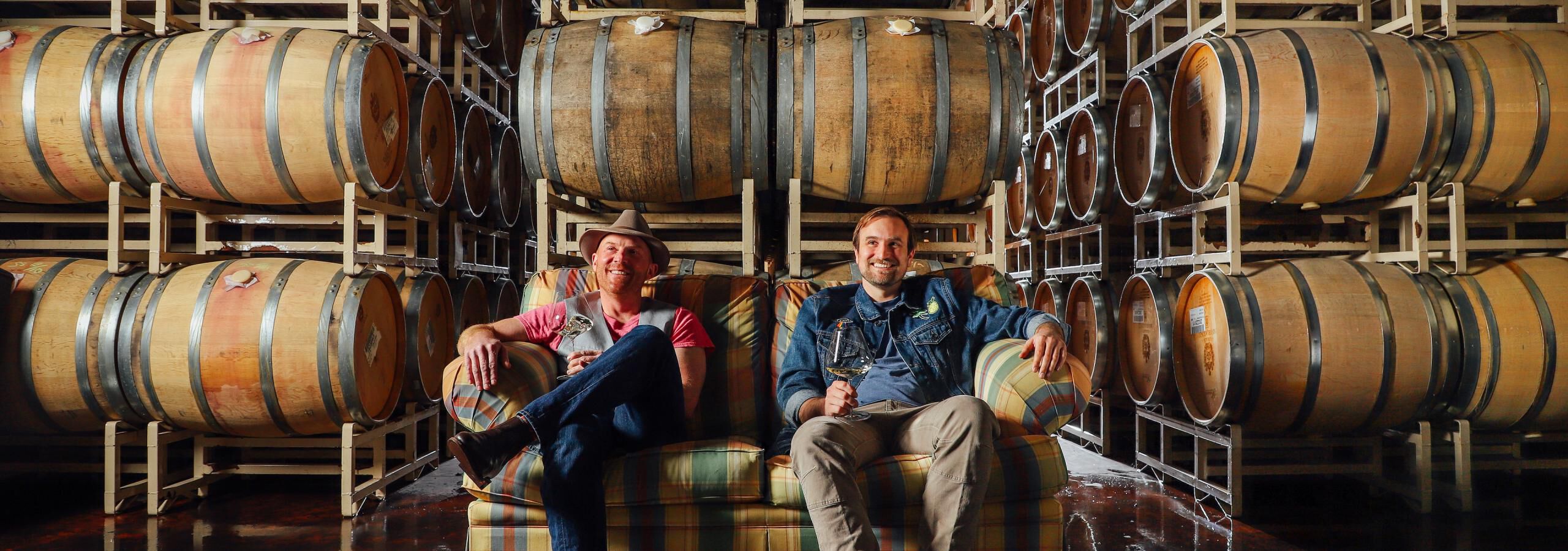 Shane and Myles holding wine glasses sitting on a plaid couch in the cellar